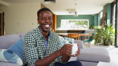 Portrait-Of-Smiling-Man-Relaxing-On-Sofa-In-Lounge-Dining-Room-At-Home-Holding-Hot-Drink