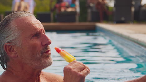 Mother-And-Son-Splashing-As-He-Eats-Ice-Lolly-At-Edge-Of-Swimming-Pool-On-Family-Summer-Holiday