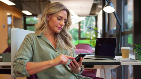 Mature-Businesswoman-Sitting-Taking-A-Break-At-Desk-In-Office-Texting-Or-Browsing-On-Mobile-Phone