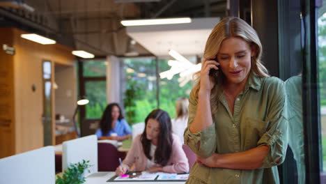 Mature-Businesswoman-Standing-By-Window-In-Busy-Office-Talking-On-Mobile-Phone