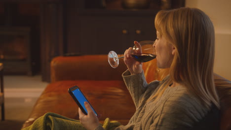 Frau-Sitzt-Zu-Hause-Auf-Dem-Sofa-Mit-Feuer-Und-Hat-Ein-Handy-Mit-Blauem-Bildschirm,-Das-Ein-Glas-Wein-Hält