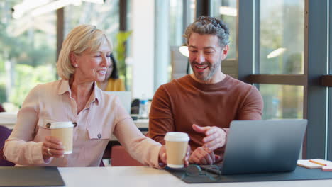 Female-Businesswoman-Brings-Takeaway-Coffee-For-Male-Colleague-As-They-Have-Meeting-With-Laptop