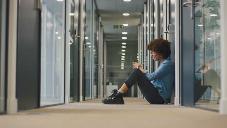Young-Businesswoman-Sitting-On-Floor-In-Corridor-Of-Modern-Office-With-Phone-Celebrating-Good-News