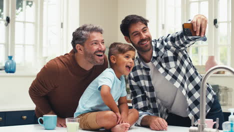 Gleichgeschlechtliche-Familie-Mit-Zwei-Vätern,-Die-In-Der-Küche-Ein-Selfie-Machen,-Während-Ihr-Sohn-Auf-Der-Theke-Sitzt
