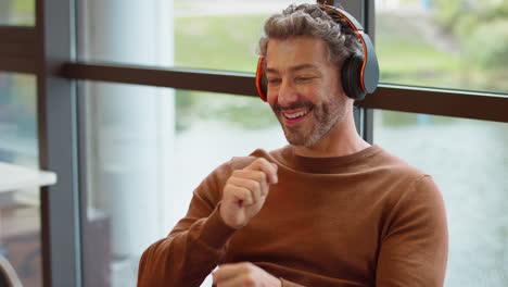 Mature-Businessman-With-Feet-On-Desk-In-Office-Listens-To-Music-On-Wireless-Headphones-Air-Drumming
