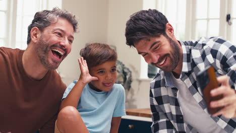 Same-Sex-Family-With-Two-Dads-Pulling-Faces-For-Selfie-In-Kitchen-With-Son-Sitting-On-Counter