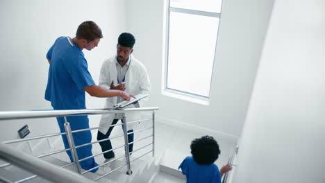 Male-Doctor-And-Nurse-With-Clipboard-Discussing-Patient-Notes-On-Stairs-In-Hospital-Building