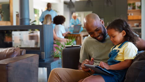 Vater-Und-Tochter-Nutzen-Zu-Hause-Ein-Digitales-Tablet,-Im-Hintergrund-Eine-Familie-Mit-Mehreren-Generationen