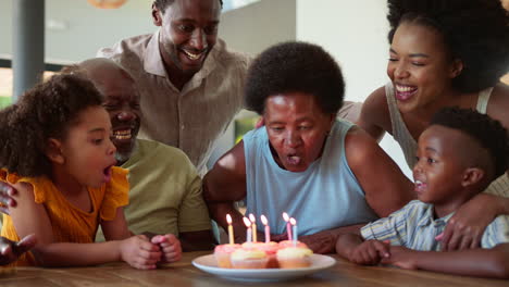 Una-Familia-Multigeneracional-Celebra-El-Cumpleaños-De-La-Abuela-Con-Pastel-Y-Velas-Alrededor-De-La-Mesa-En-Casa