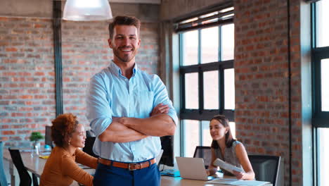 Portrait-Of-Smiling-Businessman-Standing-In-Busy-Office-With-Colleagues-Working-In-Background