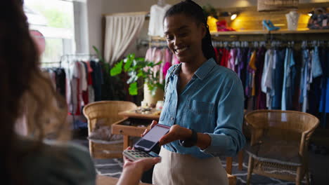 Female-Customer-In-Fashion-Store-Paying-For-Clothes-With-Contactless-Mobile-Phone-Payment-App