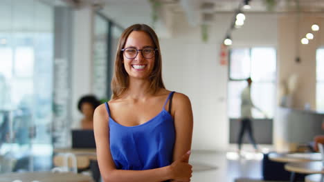 Portrait-Of-Young-Smiling-Businesswoman-Wearing-Glasses-Standing-In-Busy-Modern-Open-Plan-Office