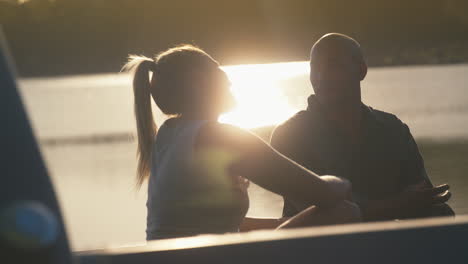 Couple-Sitting-In-Back-Of-Pick-Up-Truck-On-Road-Trip-By-Lake-At-Sunset