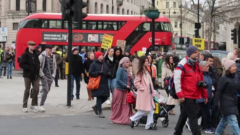 Multi-ethnic-pedestrians-crossing-street-on-crosswalk-in-London-city-with-red-bus-and-people-protesting-in-Background