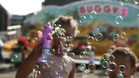 African-American-girls-blowing-soap-bubbles-with-unicorn-bubble-blower-against-fairground-background---Slow-motion---Paris,-France