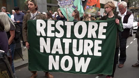 In-slow-motion-two-people-hold-a-green-banner-that-reads,-“Restore-Nature-Now”-on-a-protest-outside-the-The-Department-for-Environment-Food-and-Rural-Affairs-
