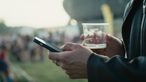 Unrecognizable-caucasian-man-browsing-phone-on-music-festival-while-drinking-beer.