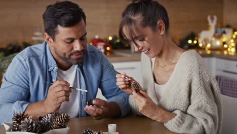 Multi-ethnicity-couple-decorating-cone-with-white-paint-during-Christmas-time-at-home.