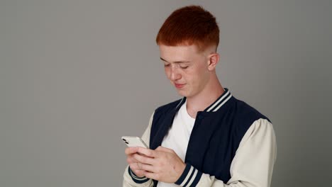 Caucasian-teenage-male-student-using-mobile-phone-on-grey-background--and-looking-at-camera.