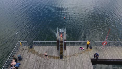 Grupo-De-Amigos-Saltando-Al-Lago-Desde-Un-Muelle-De-Madera,-Vista-Aérea