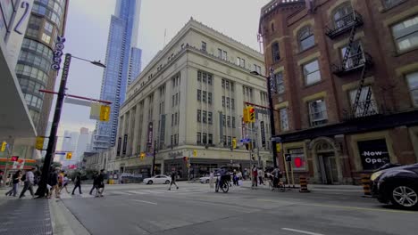 Toronto-Yonge-College-Park-Edificio-Histórico-En-El-Centro-De-La-Ciudad-En-Primer-Plano-Con-Modernos-Rascacielos-Futuristas-En-El-Fondo-De-La-Intersección-Urbana-Escena-De-La-Calle-Tráfico-Taxi-Mensajero-Motos-Personas