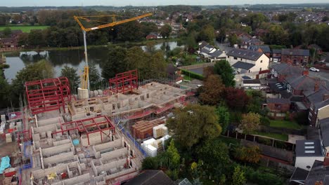 Aerial-view-looking-across-crane-on-residential-care-home-development-construction-progress-in-rural-British-village-next-to-fishing-lake