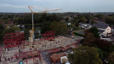 Aerial-view-rising-across-crane-building-waterside-care-home-construction-framework-in-rural-British-village-next-to-fishing-lake