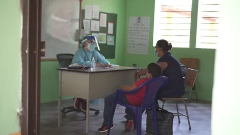 Madre-Y-Su-Hijo-Sentados-Frente-A-Un-Médico-En-Una-Consulta-Médica-Durante-Una-Brigada-De-Salud,-En-Una-Clínica-Improvisada,-En-Una-Escuela-Comunitaria-Pobre.