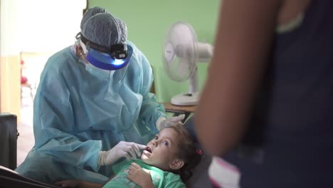 Dentist-woman-checks-the-mouth-of-a-little-girl-during-a-medical-brigade-in-an-improvised-clinic,-in-a-poor-community-school