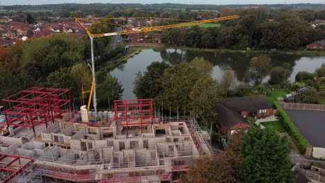 Aerial-view-crane-building-waterside-care-home-construction-framework-in-rural-British-village-next-to-fishing-lake,-pull-back-shot