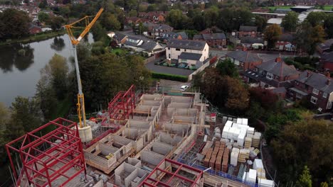 Aerial-view-circling-around-crane-on-building-waterside-care-home-construction-framework-in-rural-British-village-next-to-fishing-lake