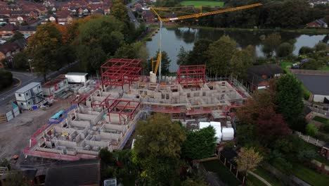 Aerial-view-orbiting-concrete-care-home-construction-framework-and-crane-in-rural-British-village-next-to-fishing-lake