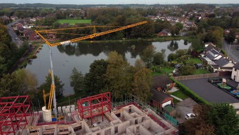 Aerial-view-circling-crane-building-waterside-care-home-construction-framework-in-rural-British-village-next-to-fishing-lake