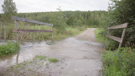 River-water-overflowing-dirt-road-bridge-in-countryside,-flooding