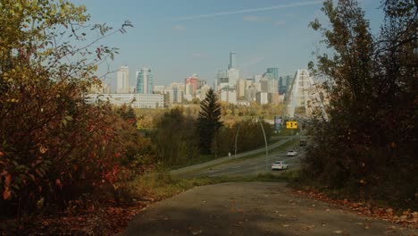 A-calm-skyline-view-of-Edmonton,-Alberta-in-the-fall-as-cars-drive-into-the-city-centre,-all-seen-through-a-peaceful-park-path-where-the-red-and-yellow-trees-begin-to-fall-from-the-branches