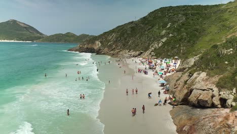 Many-tourist-in-bikini-and-bathers-at-sandy-beach-and-ocean-of-Arraial-do-Cabo-in-summer,-Brazil---Mountain-landscape-and-coastline-in-background