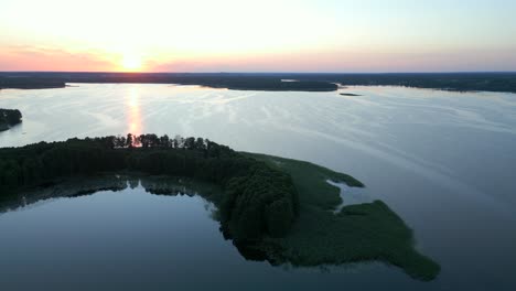 Drone-circling-over-a-small-peninsula-surrounded-by-the-calm-surface-of-a-lake,-during-sunset