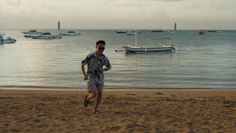 Ein-Asiatischer-Mann-In-Zeitlupe-Läuft-Bei-Sonnenuntergang-Im-Urlaub-über-Die-Kamera-Mit-Einer-Sonnenbrille-Im-Bali-Strandhintergrund