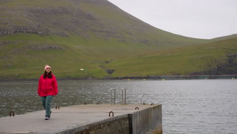 Female-traveler-walking-back-on-concrete-pier-with-view-of-volcanic-mountains-in-Faroe-Islands