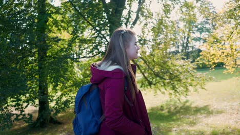 Young-girl-with-long-hair-walks-optimistically-with-a-backpack-on-her-back-in-a-sunny-park-in-autumn