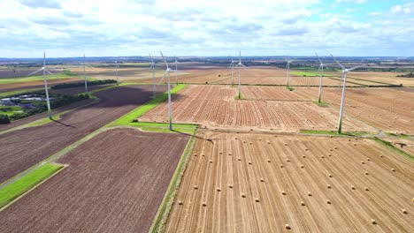 With-a-bird's-eye-view,-we-observe-the-wind-turbines-elegantly-turning-in-a-Lincolnshire-farmer's-freshly-harvested-field,-complemented-by-the-presence-of-golden-hay-bales