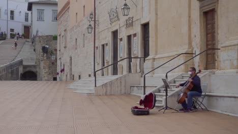 Un-Músico-Con-Una-Máscara-Tocando-El-Violonchelo-O-El-Violonchelo-En-La-Plaza-Más-Famosa-De-Spoleto-En-Umbría-Llamada-&quot;piazza-Del-Duomo&quot;