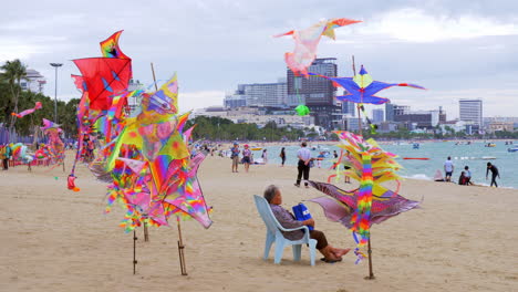 Se-Exhiben-Coloridas-Cometas-En-Postes-De-Bambú-En-Primer-Plano,-Y-En-El-Fondo-Los-Bañistas-Nadan,-Navegan-Y-Pasean-En-La-Playa-De-Pattaya,-En-La-Provincia-De-Chonburi,-En-Tailandia.