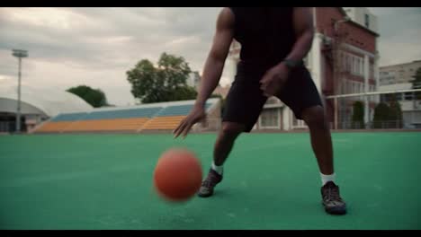 Close-up-shot-of-a-Black-skinned-basketball-player-in-black-sportswear-hitting-the-ball-off-the-floor-and-applying-tricks-to-a-stadium-in-the-city-against-a-gray-sky