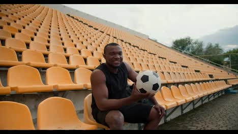 Un-Joven-Atleta-De-Piel-Negra-Se-Sienta-Con-Un-Uniforme-Deportivo-Negro-En-Las-Gradas-Amarillas-Del-Estadio-Y-Sostiene-Un-Balón-De-Fútbol-En-Sus-Manos.-Retrato-De-Un-Deportista-Feliz-Y-Exitoso