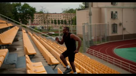 A-Black-athlete-in-a-black-sports-summer-runs-at-speed-up-the-stairs-at-the-stadium-with-yellow-chairs-in-the-stands.-Playing-sports-in-the-stands-of-the-stadium