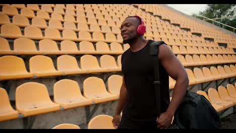 Retrato-De-Un-Atleta-De-Piel-Negra-Con-Camiseta-Negra-Y-Auriculares-Inalámbricos-Rojos,-Que-Camina-Por-El-Estadio-Con-Sillas-Amarillas-En-Las-Gradas,-Buscando-Su-Lugar