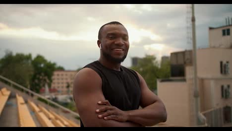 An-athlete-with-black-skin-in-a-Black-T-shirt-and-white-wireless-headphones-poses-against-the-backdrop-of-a-city-stadium-with-yellow-chairs-in-the-stands