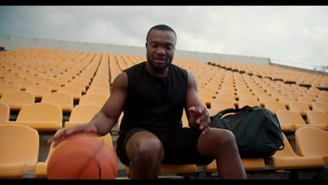 Retrato-De-Un-Deportista-Feliz-De-Piel-Negra-Con-Una-Camiseta-Negra-Que-Golpea-Una-Pelota-De-Baloncesto-Desde-El-Suelo-Y-Mira-A-La-Cámara.-Feliz-Joven-Deportista-En-Las-Gradas-Amarillas-Del-Estadio