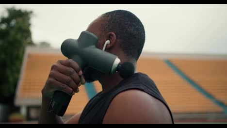 An-athlete-with-Black-skin-in-black-sportswear-after-a-workout-kneads-his-shoulders-with-a-special-massager-in-a-stadium-with-yellow-stands